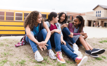 A group of four middle school girls laughing on the lawn in front of a school bus