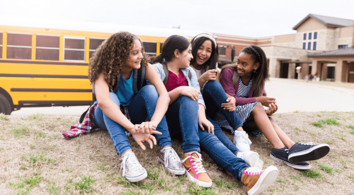 A group of four middle school girls laughing on the lawn in front of a school bus