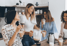 4 women friends smiling and making drinks together in a kitchen