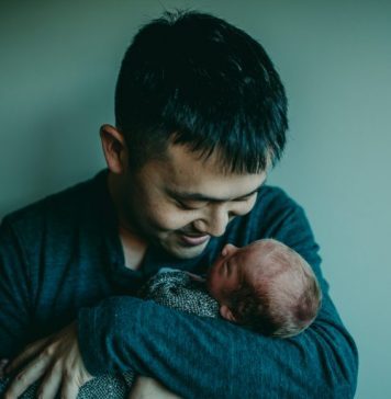 A father holding and staring at his newborn baby with a smile on his face in front of a teal background