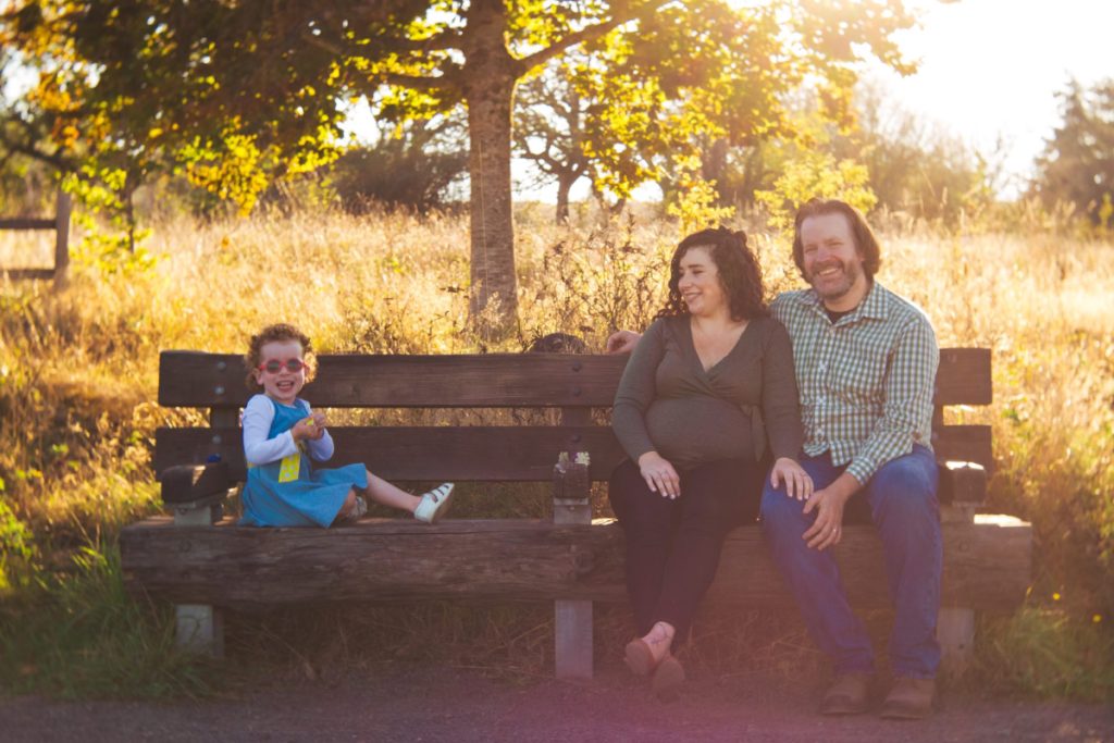 A family of three with a small child sit on a bench smiling at Powell Butte in the Fall
