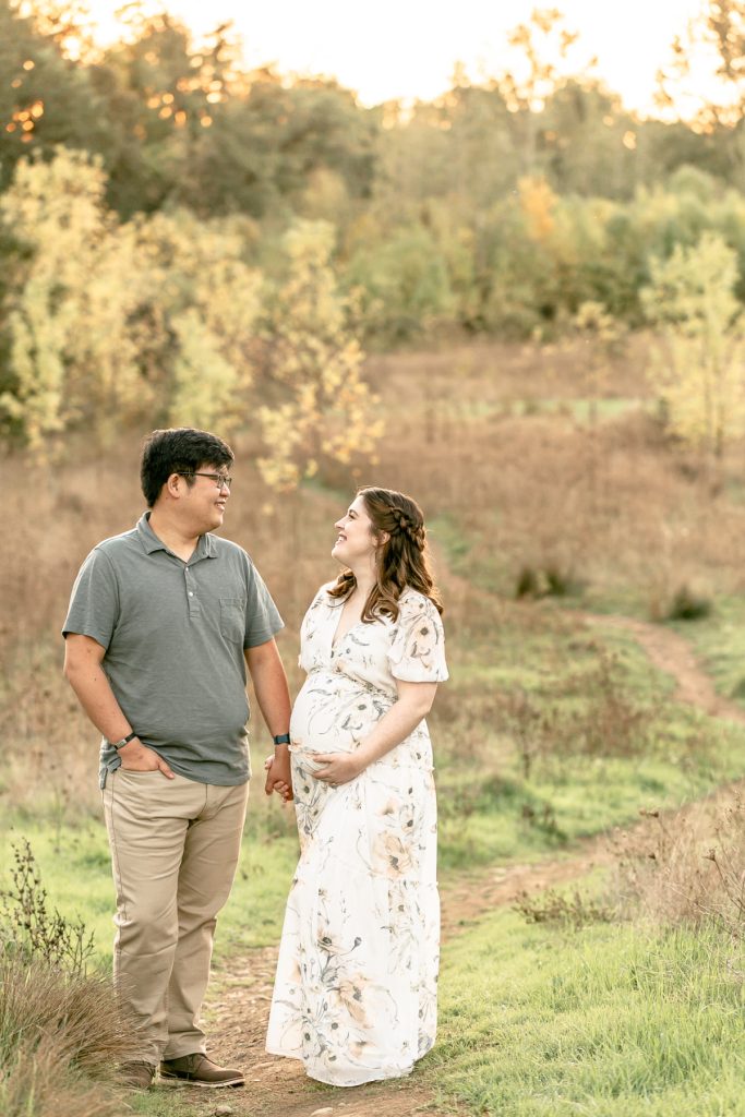 A pregnant woman and a man holding hands and smiling in front of some trees at Powell Butte