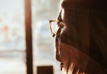 Woman in glasses looks pensively out a window while the sun shines in