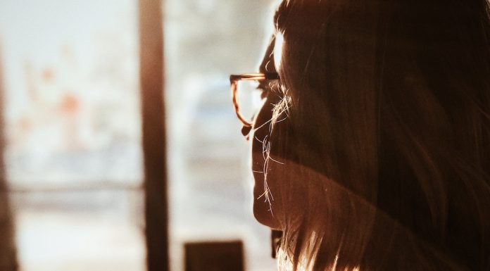 Woman in glasses looks pensively out a window while the sun shines in