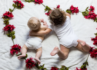 Two babies lie on a blanked together, surrounded by a heart made of flowers an d leaves