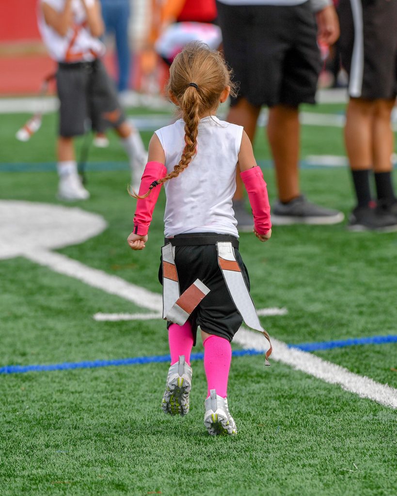 Image of young girl walking onto the football field with her flags attached for a flag football game.